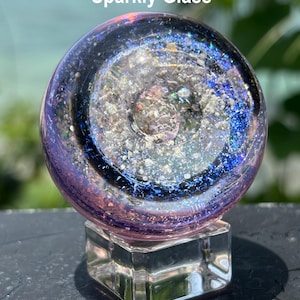 Pode incluir: Uma esfera de vidro dicr&oacute;ico com um design brilhante, semelhante a uma gal&aacute;xia, repousando sobre uma base de vidro transparente e quadrada. A esfera apresenta tons de roxo, azul e prata, com o texto "Add Dichroic Glass 'Sparkly Glass'".