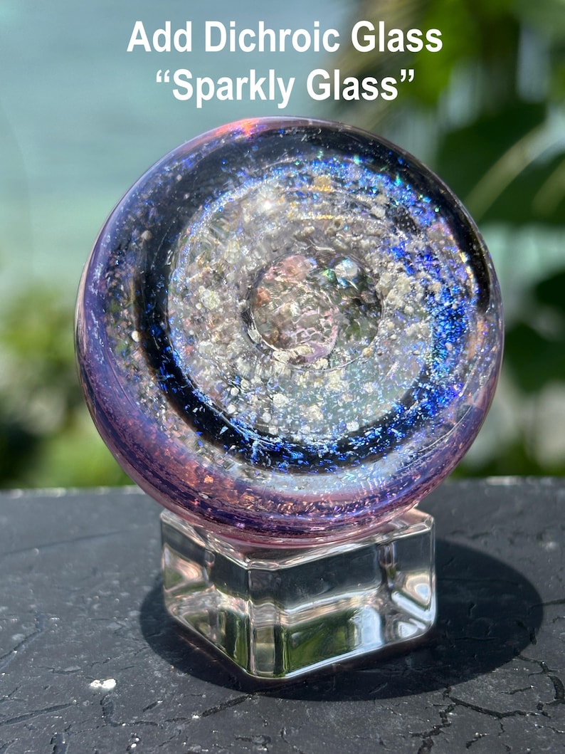 Pode incluir: Uma esfera de vidro dicr&oacute;ico com um design brilhante, semelhante a uma gal&aacute;xia, repousando sobre uma base de vidro transparente e quadrada. A esfera apresenta tons de roxo, azul e prata, com o texto "Add Dichroic Glass 'Sparkly Glass'".