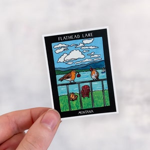 May include: A rectangular sticker featuring a scenic illustration of Flathead Lake in Montana. The artwork depicts two birds perched on a railing, a lake, mountains, and a sailboat. The sticker has a black border and the words "FLATHEAD LAKE" and "MONTANA".