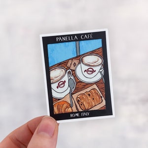 May include: A sticker featuring a cartoon illustration of two cups of coffee and pastries on a table. The sticker says "Panella Cafe" and "Rome Italy".