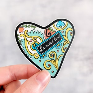 May include: A heart-shaped sticker with a blue, yellow, and white paisley pattern. The sticker has a black border and a small black rectangle with the text "La vita è bella" in white.