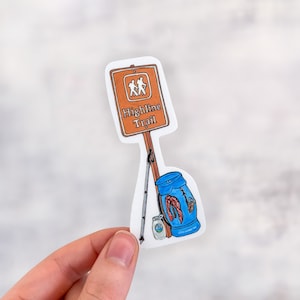 May include: A sticker featuring a brown sign with the words "Highline Trail" and a graphic of two hikers. A blue backpack, a jar, and a walking stick are also depicted. The sticker is held by a hand.