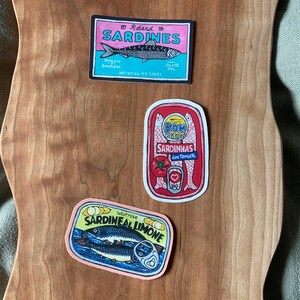 Puede incluir: Tres etiquetas de latas de sardinas de estilo vintage. La etiqueta superior es rosa y blanca con el texto "Roland Sardines" y un dibujo de sardinas en aceite de oliva. La etiqueta del medio es roja y blanca con el texto "Bom Petiscos Sardinhas em Tomate" y un dibujo de sardinas en salsa de tomate. La etiqueta inferior es amarilla y rosa con el texto "Waitrose Sardineal Limone" y un dibujo de sardinas en salsa de lim&oacute;n.