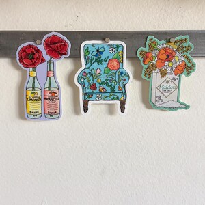 Pode incluir: Tr&ecirc;s adesivos com designs florais. O primeiro adesivo mostra duas garrafas de refrigerante italiano com flores vermelhas. O segundo adesivo mostra uma poltrona azul com um padr&atilde;o floral. O terceiro adesivo mostra uma caixa de flocos de sal marinho Maldon com um buqu&ecirc; de flores vermelhas e brancas.