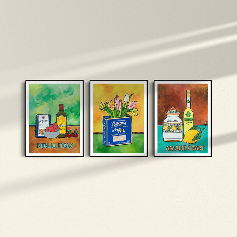 A triptych of colorful framed artworks on a light wall. The first shows Italian ingredients labeled Puglia, Italy. The middle depicts a Ronzoni pasta box with tulips. The third features limoncello and lemons labeled Amalfi Coast.