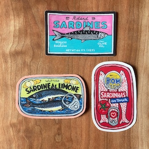 Puede incluir: Tres etiquetas de latas de sardinas de colores. La primera etiqueta es azul, rosa y blanca con el texto "Roland Sardines" y un dibujo de una sardina. La segunda etiqueta es rosa, amarilla y blanca con el texto "Waitrose Sardineal Limone" y un dibujo de dos sardinas. La tercera etiqueta es roja, blanca y amarilla con el texto "Bom Reitisco Sardinhas em tomate" y un dibujo de tres sardinas.