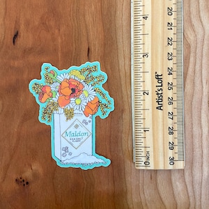 Pode incluir: Um adesivo de um saleiro com um buqu&ecirc; de flores na frente. O saleiro &eacute; branco com detalhes azuis e diz "Maldon Sea Salt Flakes". As flores s&atilde;o laranja, amarelo e branco.