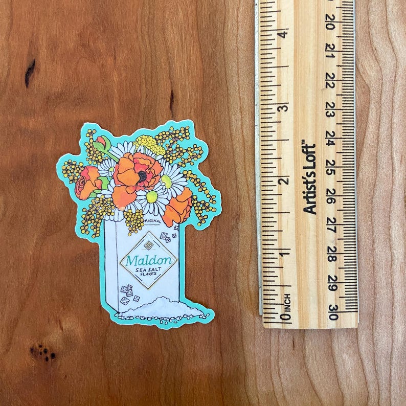 Pode incluir: Um adesivo de um saleiro com um buqu&ecirc; de flores na frente. O saleiro &eacute; branco com detalhes azuis e diz "Maldon Sea Salt Flakes". As flores s&atilde;o laranja, amarelo e branco.