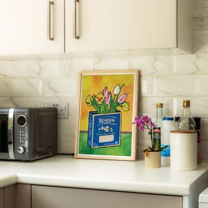 A colorful framed artwork featuring a blue Ronzoni pasta box filled with tulips, displayed on a kitchen countertop. The countertop also holds a microwave, an orchid plant, a white canister, and bottles of vinegar and other condiments.