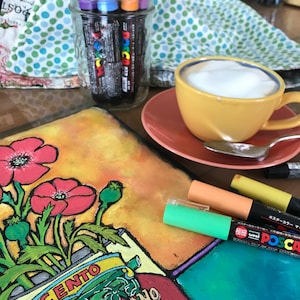 Pode incluir: Um close-up de uma pintura em tela com papoulas vermelhas e folhas verdes. A pintura est&aacute; sobre uma mesa com uma x&iacute;cara de caf&eacute;, uma colher e v&aacute;rios marcadores. Os marcadores est&atilde;o em um frasco de vidro e s&atilde;o rotulados como "Posca" e "Uni Posca".