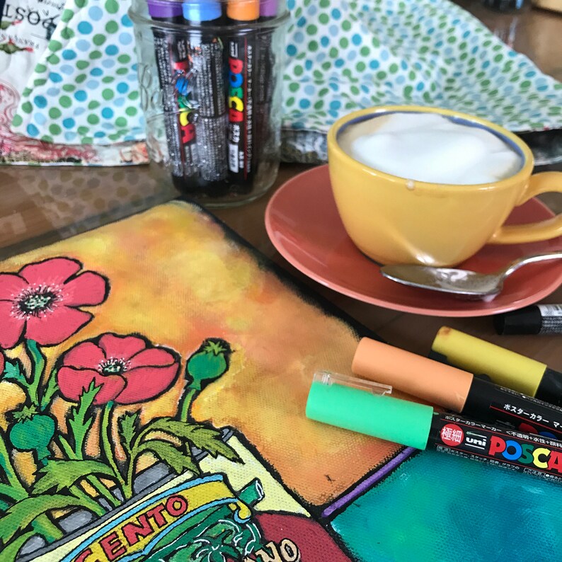 Pode incluir: Um close-up de uma pintura em tela com papoulas vermelhas e folhas verdes. A pintura est&aacute; sobre uma mesa com uma x&iacute;cara de caf&eacute;, uma colher e v&aacute;rios marcadores. Os marcadores est&atilde;o em um frasco de vidro e s&atilde;o rotulados como "Posca" e "Uni Posca".