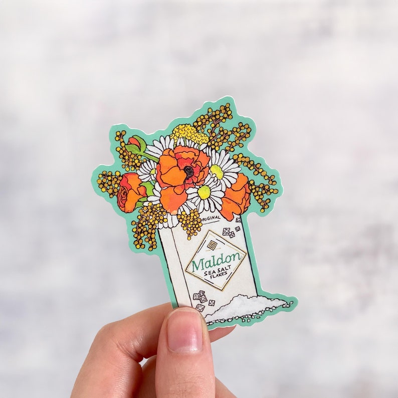 Pode incluir: Um adesivo com um buqu&ecirc; de flores laranja e brancas em um pequeno recipiente com o texto "Maldon Sea Salt Flakes" na frente.
