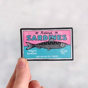Puede incluir: Una ilustraci&oacute;n en blanco y negro de una etiqueta de lata de sardinas. La etiqueta es rosa y azul con el texto "Est. Roland 1918 Sardines Skinless & Boneless - In Olive Oil Net Wt 4.4 oz (125g)"