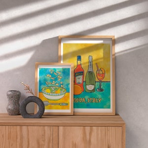 May include: Two framed watercolour illustrations. The first shows a bowl of pasta with a yellow background. The second shows a bottle of Aperol, a bottle of prosecco, and a glass of Aperol spritz with a blue and yellow background. The text "Adua, Italy" is below the illustration.