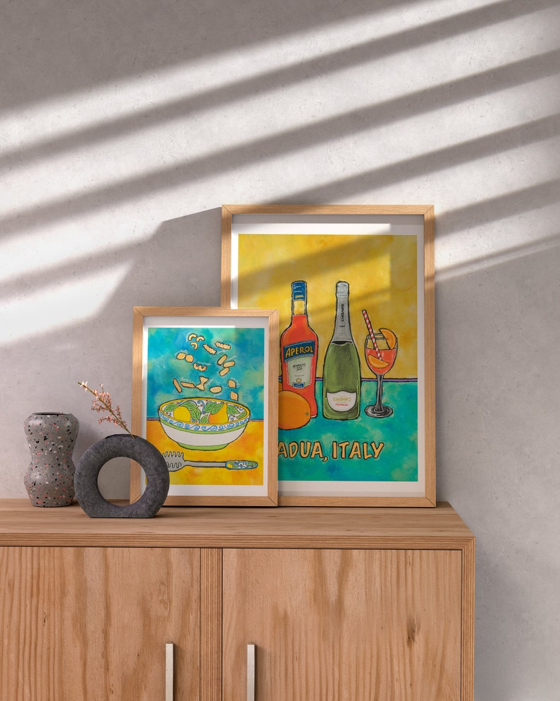 May include: Two framed watercolour illustrations. The first shows a bowl of pasta with a yellow background. The second shows a bottle of Aperol, a bottle of prosecco, and a glass of Aperol spritz with a blue and yellow background. The text "Adua, Italy" is below the illustration.
