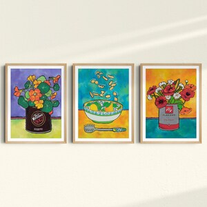 May include: Three framed art prints in a colourful, whimsical style. The first print features a can of beans with a bouquet of orange flowers. The second print shows a bowl of soup with pasta falling from the sky. The third print features a can of soup with a bouquet of red flowers.
