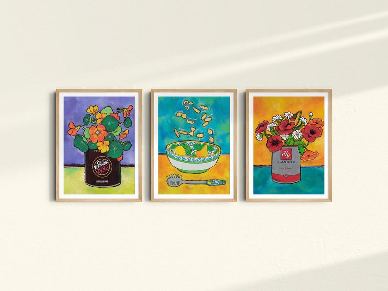 May include: Three framed art prints in a colourful, whimsical style. The first print features a can of beans with a bouquet of orange flowers. The second print shows a bowl of soup with pasta falling from the sky. The third print features a can of soup with a bouquet of red flowers.