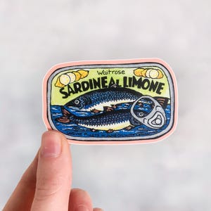 Puede incluir: Una pegatina rosa y amarilla con una ilustraci&oacute;n de dibujos animados de una lata de sardinas. La lata dice "Waitrose Sardine al Limone" y "Sardine fillets in olive oil with lemon".