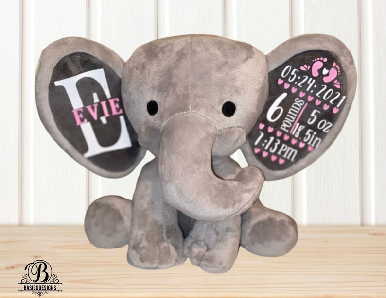 Puede incluir: Un elefante de peluche gris con ojos negros y una trompa. Las orejas del elefante tienen texto personalizado: una oreja dice "EVIE" y la otra oreja tiene los detalles del nacimiento: 05.24.2021, 2,7 kg, 142 g, 47 cm, y 19:13. Un regalo para beb&eacute;.