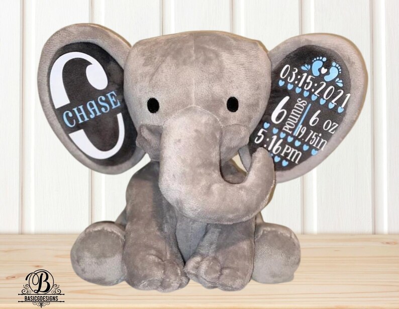 Puede incluir: Un elefante de peluche gris con orejas grandes. La oreja izquierda muestra el nombre "CHASE" en azul. La oreja derecha muestra detalles de nacimiento: 15/03/2021, 2,72 kg, 49,53 cm y 17:16, en azul. Un art&iacute;culo tem&aacute;tico para beb&eacute;s.