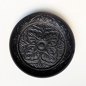 May include: Black circular incense burner with an intricate floral design. The burner has a small hole in the center for burning incense.