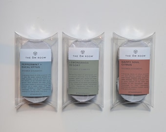 May include: Three shower steamers in clear plastic pillow boxes. The left box has a blue label reading THE OM ROOM PEPPERMINT + EUCALYPTUS. The center box has a green label reading THE OM ROOM LEMONGRASS RESORT. The right box has a pink label reading THE OM ROOM HAPPY SOUL CITRUS.