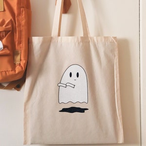 May include: A white canvas tote bag with a black and white illustration of a ghost with a friendly face. The ghost is holding its arms out in a welcoming gesture.