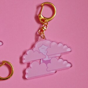 May include: A pink and white acrylic keychain featuring a cartoon woman with closed eyes inside two clouds. The keychain has a gold metal ring and chain.