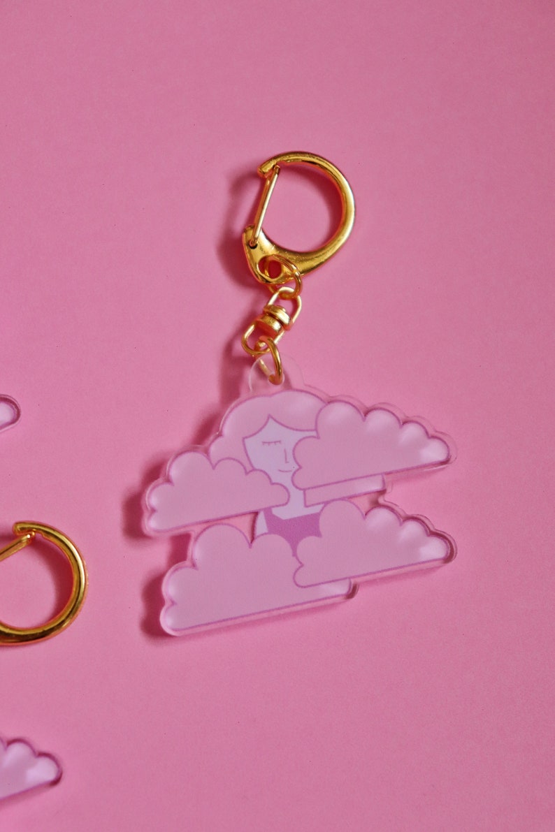 May include: A pink and white acrylic keychain featuring a cartoon woman with closed eyes inside two clouds. The keychain has a gold metal ring and chain.