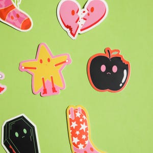 May include: A set of colorful stickers featuring various characters and objects, including a broken heart, a star, an apple, a cowboy boot, and a coffin. The stickers are on a green background.