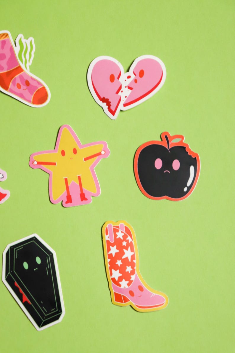 May include: A set of colorful stickers featuring various characters and objects, including a broken heart, a star, an apple, a cowboy boot, and a coffin. The stickers are on a green background.