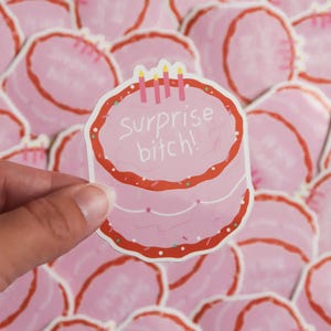 May include: A pink sticker in the shape of a cake with red frosting and three candles. The text "surprise bitch!" is written on the cake. The sticker is held in a hand, with other similar stickers in the background.