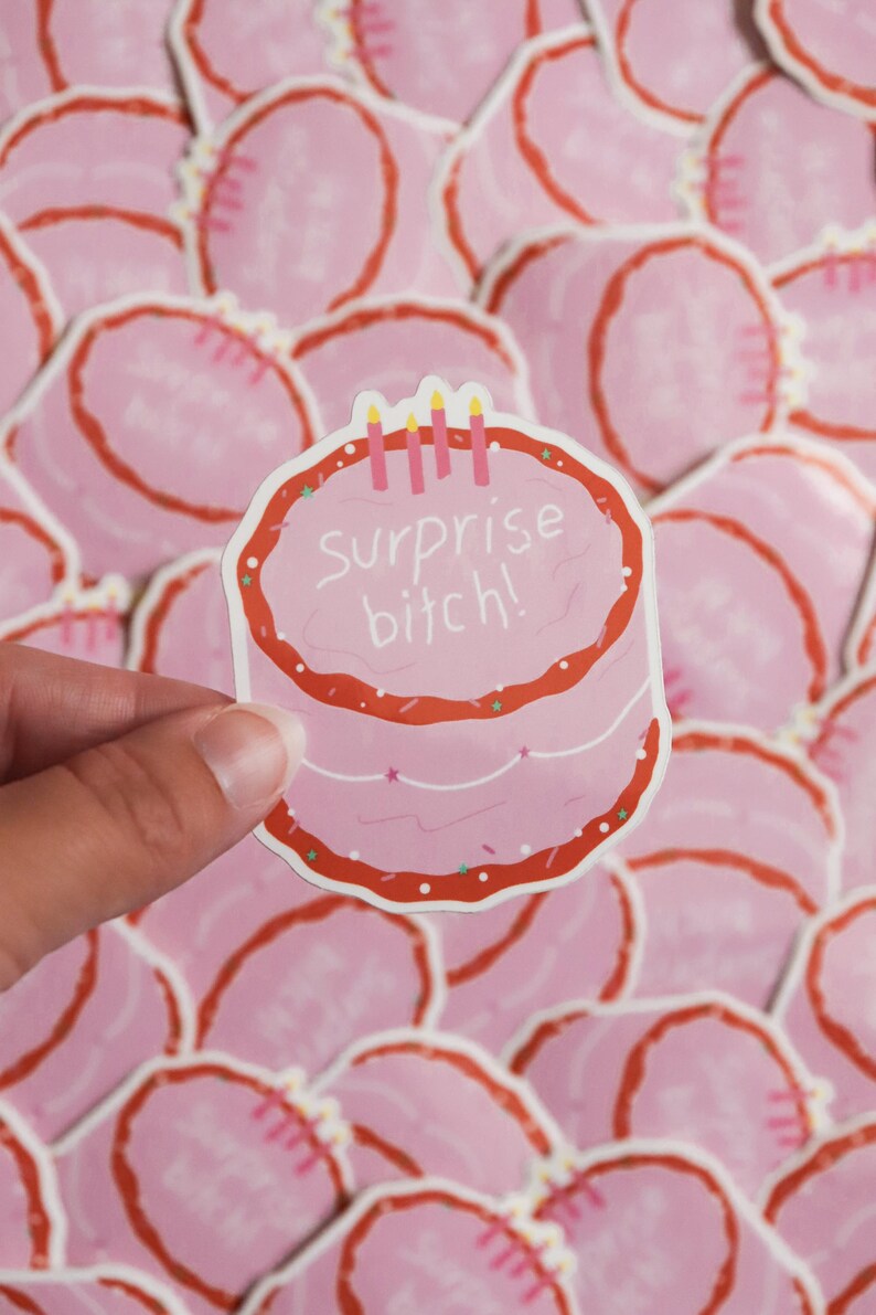 May include: A pink sticker in the shape of a cake with red frosting and three candles. The text "surprise bitch!" is written on the cake. The sticker is held in a hand, with other similar stickers in the background.