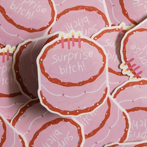 May include: Pink stickers featuring a cartoon cake design with red frosting and candles. The text "Surprise bitch!" is written on the cake. The stickers are scattered across the image.