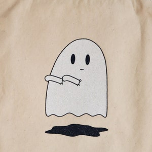 May include: A beige tote bag with a white ghost with black eyes and a smile. The ghost is holding its arms out in front of it. The ghost is outlined in black and has glitter on it.