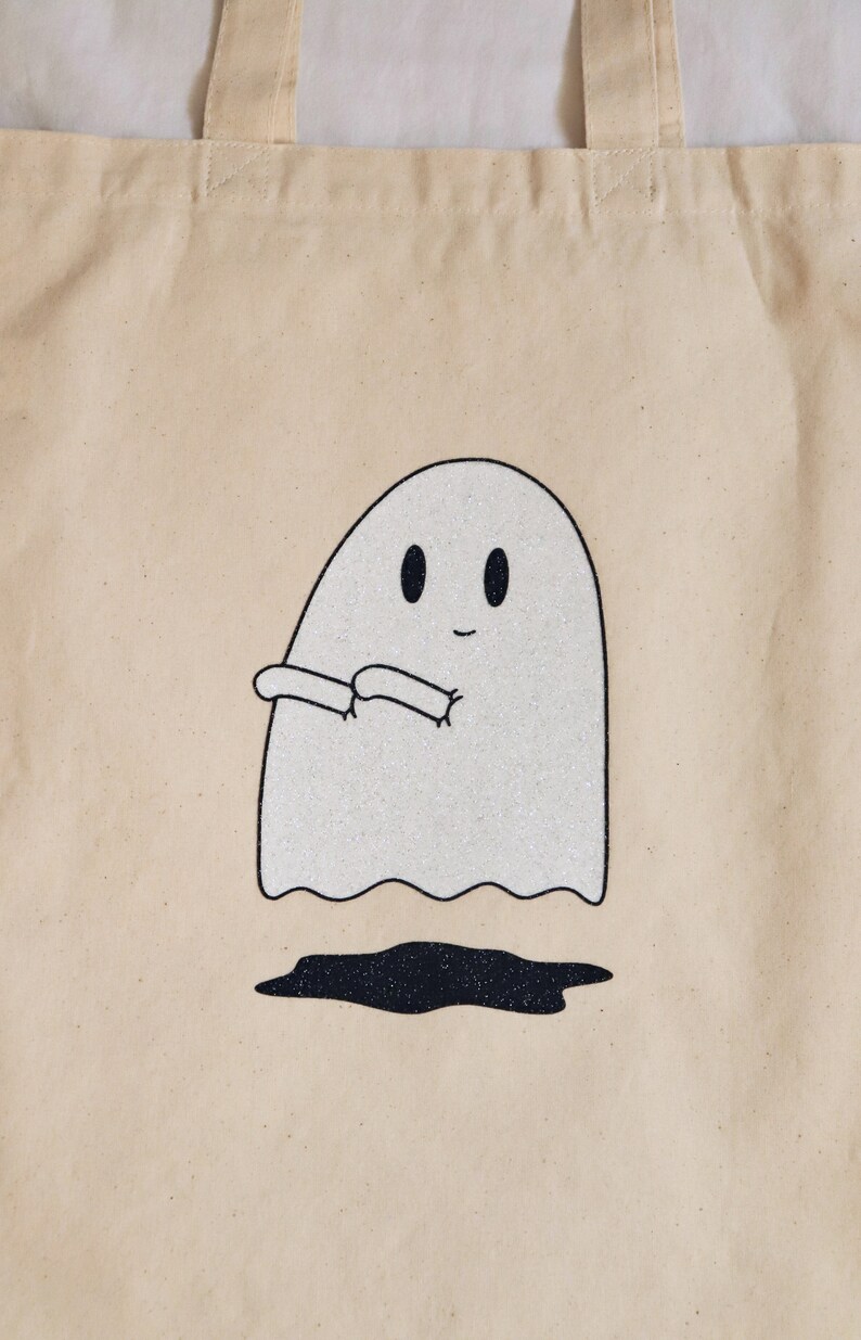 May include: A beige tote bag with a white ghost with black eyes and a smile. The ghost is holding its arms out in front of it. The ghost is outlined in black and has glitter on it.