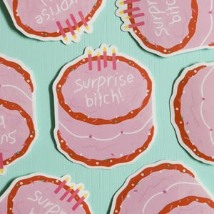 May include: Pink cake stickers with red frosting and the text "Surprise bitch!" are scattered on a light blue background. Each sticker has candles and sprinkles. The stickers are a fun, celebratory design.