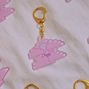 May include: A pink acrylic keychain featuring a cartoon character with closed eyes and a smile, floating on two pink clouds. The keychain has a gold chain and a gold ring.