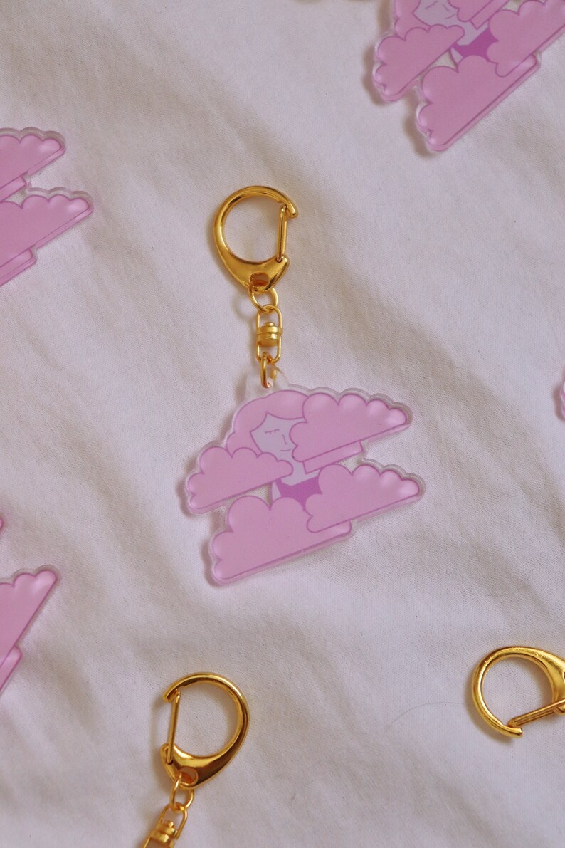 May include: A pink acrylic keychain featuring a cartoon character with closed eyes and a smile, floating on two pink clouds. The keychain has a gold chain and a gold ring.