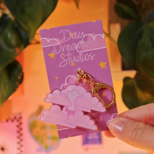 May include: Pink and gold keychain featuring a cartoon person sleeping in a cloud with the text "Daydream Studios" and "Thank you!" on a pink background with gold stars.