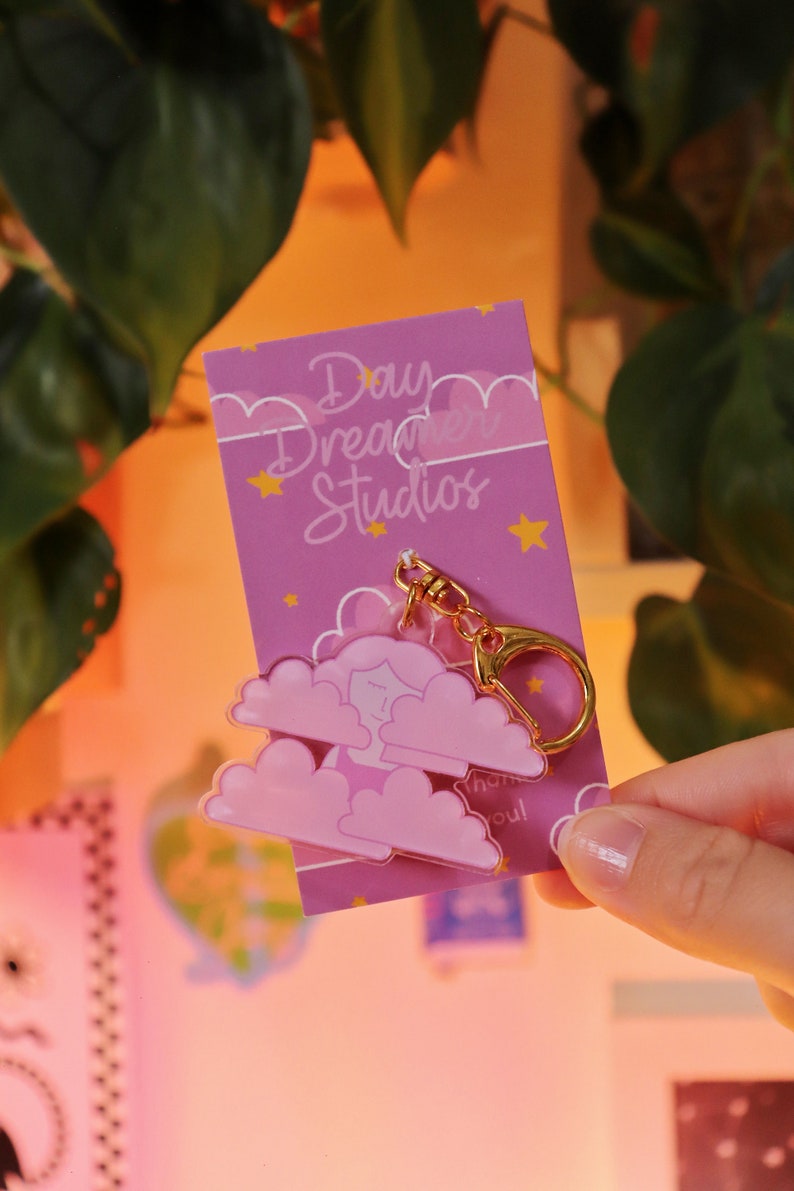 May include: Pink and gold keychain featuring a cartoon person sleeping in a cloud with the text "Daydream Studios" and "Thank you!" on a pink background with gold stars.