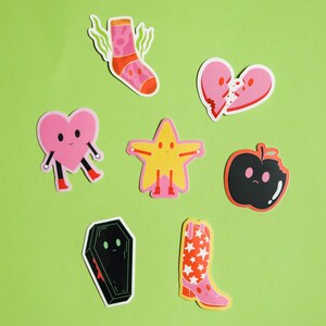 May include: A set of seven colorful stickers featuring various cartoon characters. The stickers include a pink heart, a yellow star, a black apple, a pink sock, a black coffin, a broken pink heart, and a pink cowboy boot with red stars.
