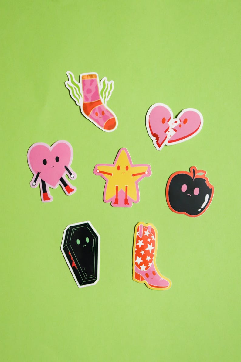 May include: A set of seven colorful stickers featuring various cartoon characters. The stickers include a pink heart, a yellow star, a black apple, a pink sock, a black coffin, a broken pink heart, and a pink cowboy boot with red stars.