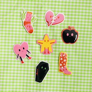 May include: A set of eight colorful stickers featuring various cartoon characters. The stickers include a sock, a broken heart, an apple, a star, a coffin, and a cowboy boot. The stickers are all on a green and white checkered background.