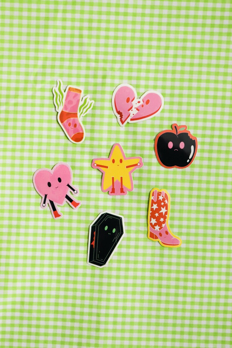 May include: A set of eight colorful stickers featuring various cartoon characters. The stickers include a sock, a broken heart, an apple, a star, a coffin, and a cowboy boot. The stickers are all on a green and white checkered background.