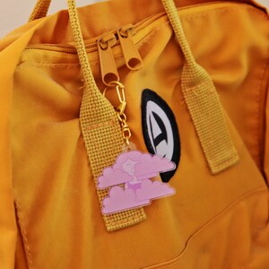 May include: A yellow backpack with a zipper closure and a pink cloud keychain attached to the zipper pull. The keychain features a small, cartoon-like face in the center of the cloud.