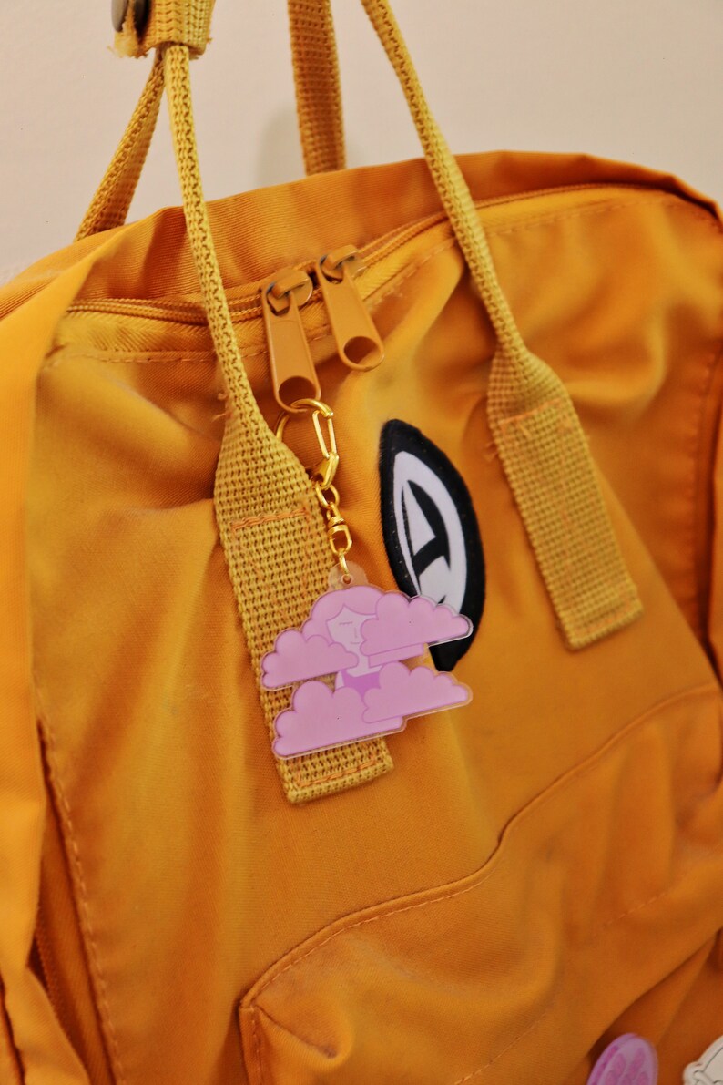May include: A yellow backpack with a zipper closure and a pink cloud keychain attached to the zipper pull. The keychain features a small, cartoon-like face in the center of the cloud.