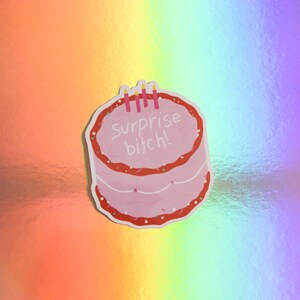 May include: A pink sticker in the shape of a cake with red frosting and the words "Surprise bitch!" in white. The sticker has three pink candles on top. The background is a holographic rainbow pattern.