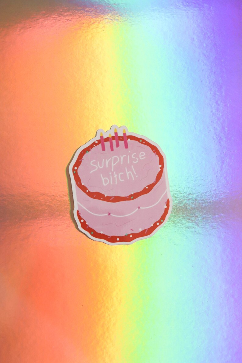 May include: A pink sticker in the shape of a cake with red frosting and the words "Surprise bitch!" in white. The sticker has three pink candles on top. The background is a holographic rainbow pattern.