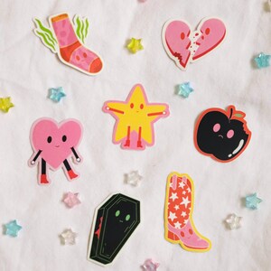 May include: A set of eight colorful stickers featuring various characters, including a sock, a broken heart, a star, an apple, a heart, a coffin, and a cowboy boot. The stickers are on a white background.